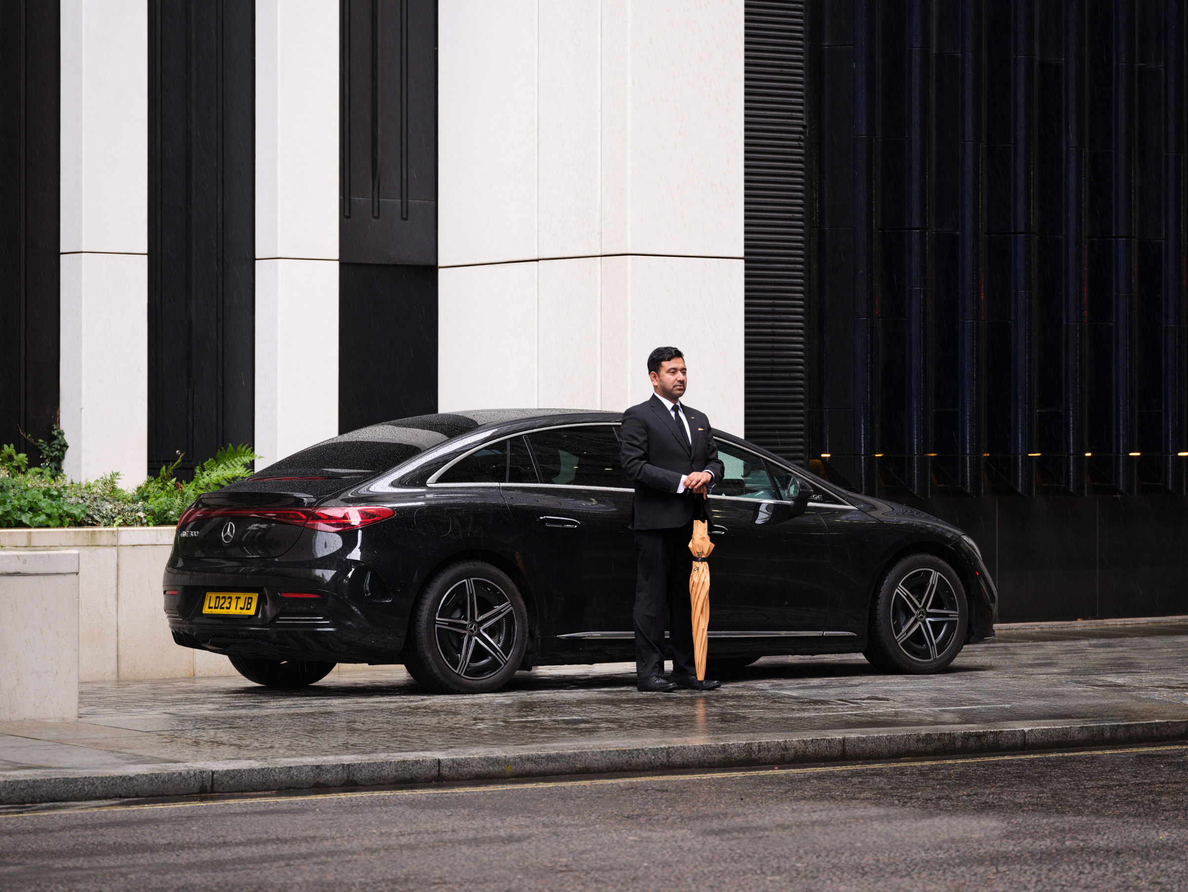 Professional chauffeur in a suit standing next to a black luxury Business Class vehicle.